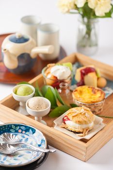 Dessert set for tea time with cake, ice cream, egg tart, and scone in wood tray on white table with tea set and diashes