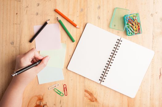 Notebook and sticky paper, paper clip, color pencil, and pen with female left hand on wood table