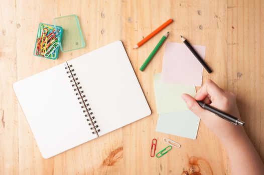 Notebook and sticky paper, paper clip, color pencil, and pen with female right hand on wood table