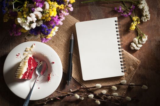 Small notepad with pen, pencil and bits of red velvet cake on rustic wood background with low key scene.