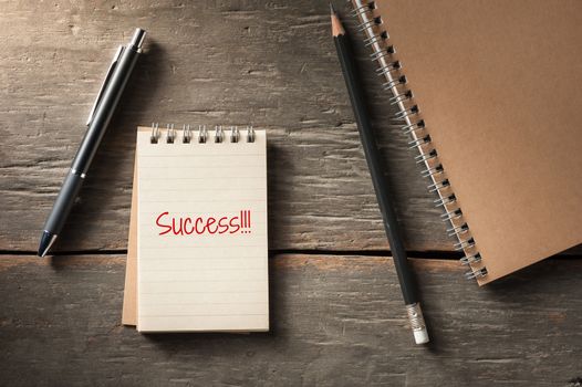 Small notepad with the word success, pen, and pencil on rustic wood background with low key scene.