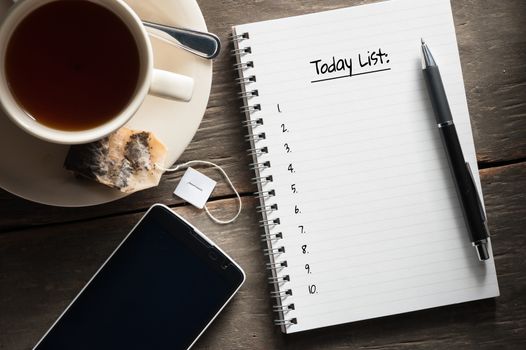 Small notepad with written messeage,  a cup of tea, pen, pencil and cellphne on rustic wood background with low key scene