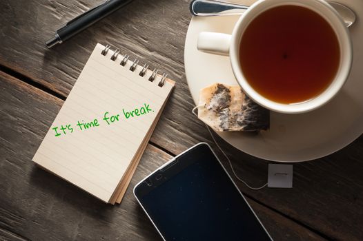 Small notepad with written messeage,  a cup of tea, pen, pencil and cellphne on rustic wood background with low key scene