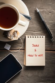 Small notepad with written words a cup of tea, pen, pencil and cellphne on rustic wood background with low key scene