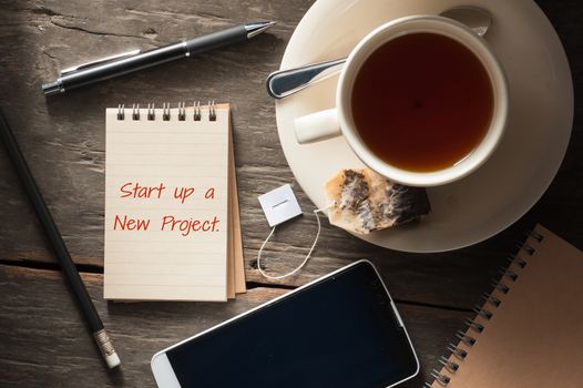 "start up a new project" is written on small notepad with a cup of tea, pen, pencil and cellphne on rustic wood background with low key scene