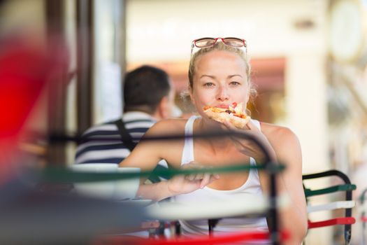 Casual blond lady eating pizza slice outdoor in typical italian street restaurant on hot summer day. Traditional italian fast food eatery.