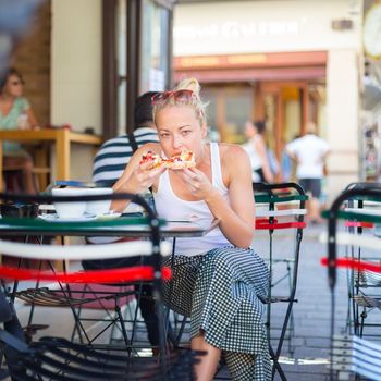 Casual blond lady eating pizza slice outdoor in typical italian street restaurant on hot summer day. Traditional italian fast food eatery.