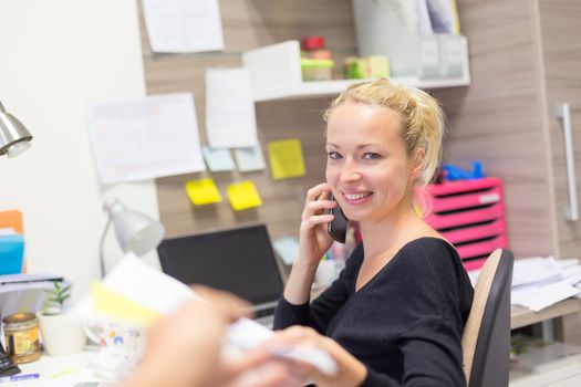 Business and entrepreneurship consept. Beautiful blonde business woman talking on mobile phone in colorful modern creative working environment accepting papers.  Female multi tasking.