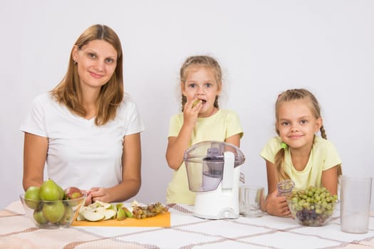 Young beautiful mother and two daughters sitting at a table squeezed juice from pears and grapes with a juicer