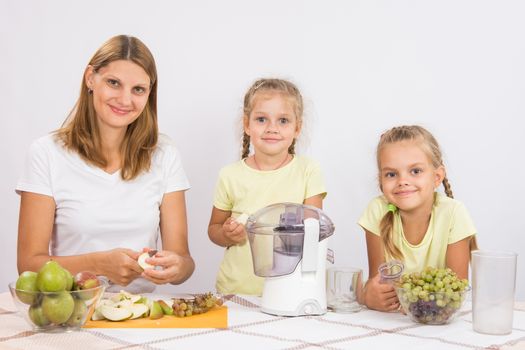 Young beautiful mother and two daughters sitting at a table squeezed juice from pears and grapes with a juicer