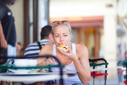 Casual blond lady eating pizza slice outdoor in typical italian street restaurant on hot summer day. Traditional italian fast food eatery.