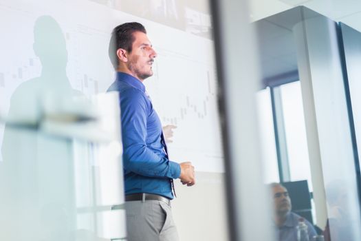 Business man making a presentation in front of whiteboard. Business executive delivering a presentation to his colleagues during meeting or in-house business training. View through glass.