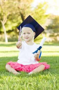 Cute Little Girl In Grass Wearing Graduation Cap Holding Diploma With Ribbon.