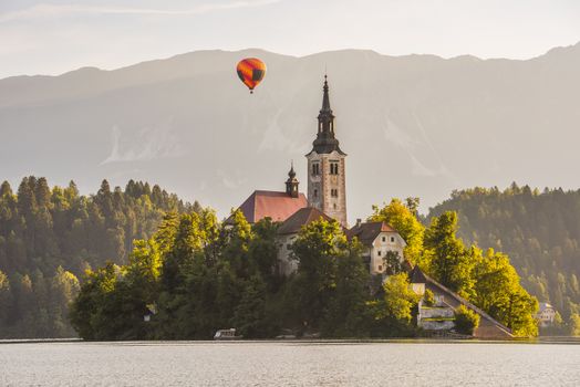Detail of Famous Catholic Church on Little Island in Bled Lake, Slovenia with Hot Air Balloon Flying with Mountains in Background at Sunrise