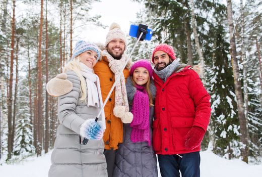 technology, season, friendship and people concept - group of smiling men and women taking selfie with smartphone and monopod in winter forest