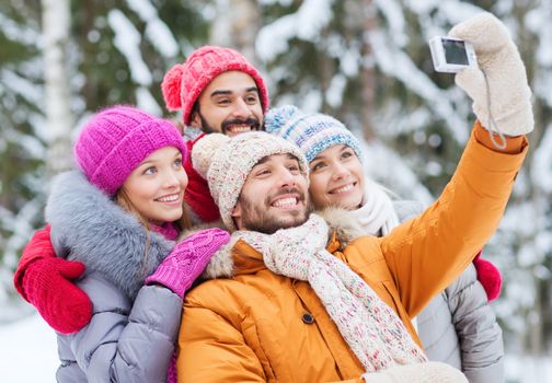 technology, season, friendship and people concept - group of smiling men and women taking selfie with digital camera in winter forest