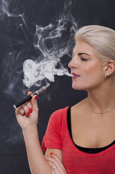 Stylish blond woman smoking an e-cigarette exhaling a cloud of smoke with her eyes closed in enjoyment, profile view on a dark background