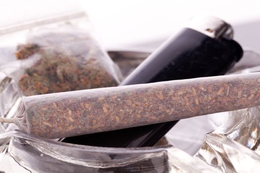 Close up of marijuana joint made with translucent rolling papers, plastic baggy of dried marijuana, black lighter and pipe on white background