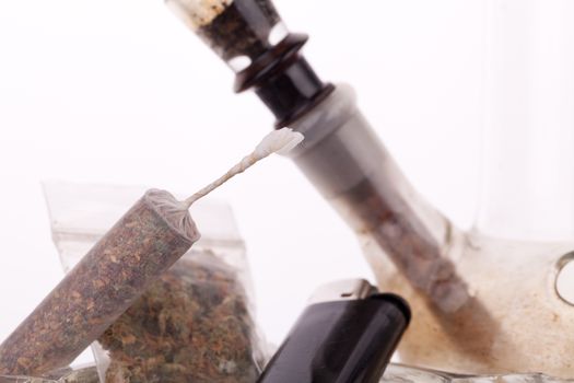 Close up of marijuana joint made with translucent rolling papers, plastic baggy of dried marijuana, black lighter and pipe on white background