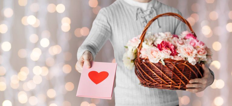 holidays, people, feelings and greetings concept - close up of man holding basket full of flowers and giving postcard over holidays lights background