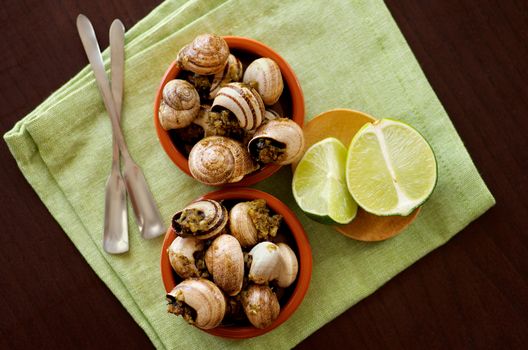 Delicious Escargot with Garlic Butter in Two Bowls with Silver Forks and Sliced Lime closeup on Green Napkin. Top View