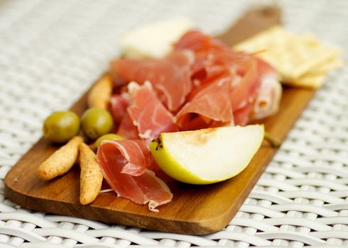 Arrangement of Spanish Tapas with Hamon, Bread Sticks, Pear and Green Olives on Wooden Cutting Board on Wicker background. Focus on Foreground