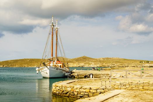 A beautiful white hulled classic Greek sailboat is tied to the stone jetty of Delos, Greece, under a powerful sky.