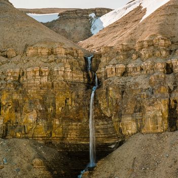 A large, high waterfall in the Arctic framed by two rugged, brown stone cliffs.