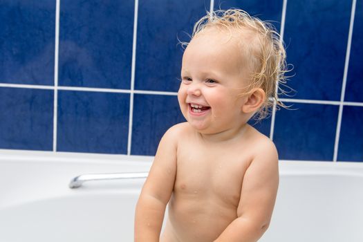 Adorable baby girl having bath in a white bathtub, smiling and looking at parent. Blue tiles behind