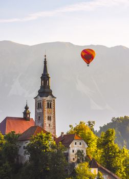 Detail of Famous Catholic Church in Bled Lake, Slovenia with Hot Air Balloon Flying with Mountains in Background at Sunrise