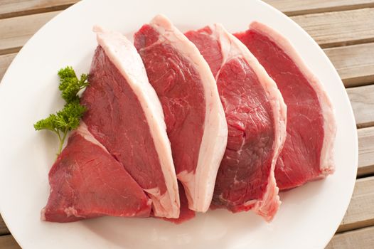 Four fresh raw beef steaks with a fatty rind displayed fanned on a plate with parsley garnish ready for cooking, overhead view