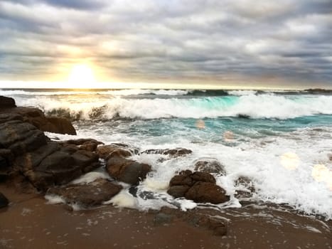 crashing blue waves over rocky and sandy beach under cloudy or overcast sky in sunset