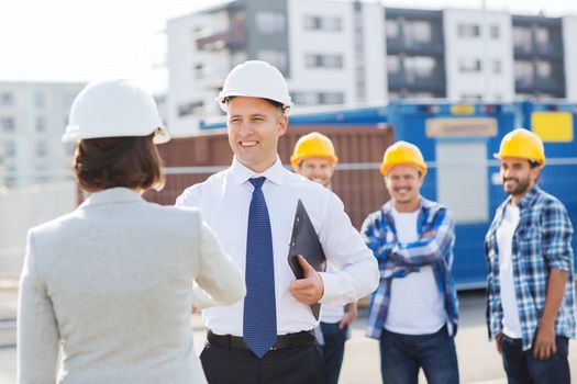 business, building, teamwork, gesture and people concept - group of smiling builders in hardhats with clipboard greeting each other outdoors