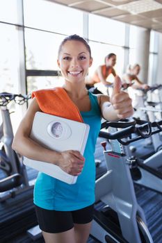 sport, fitness, slimming and people concept - smiling woman with scales and towel showing thumbs up in gym