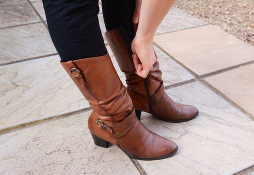 Young woman zips up her fashionable brown leather boots, standing on stone slabs