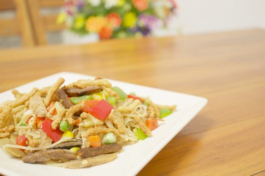 Colorful long life noodle fried with cauliflower, carrots, mushroom, red and green bell pepper and vegan protein dry vegetarian food have wooden table and chair with blur flower as background.