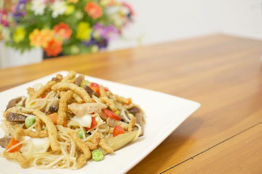Colorful long life noodle fried with cauliflower, carrots, mushroom, red and green bell pepper and vegan protein dry vegetarian food on wooden table with blur flower as background.