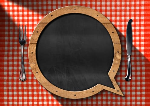 Empty blackboard in the shape of speech bubble on a table with red and white checkered tablecloth and silver cutlery. Template for a restaurant menu