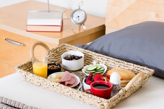 Close Up of Breakfast Tray Filled with Variety of Foods Sitting on Unmade Bed Next to Bedside Table with Lamp and Alarm Clock in Hotel Room