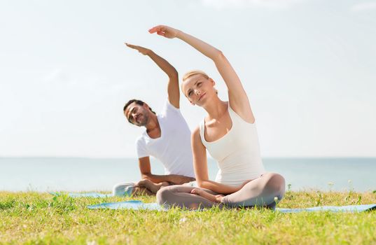 fitness, sport, friendship and lifestyle concept - smiling couple making yoga exercises sitting on mats outdoors