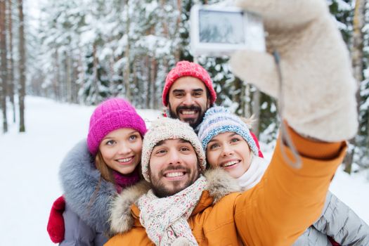 technology, season, friendship and people concept - group of smiling men and women taking selfie with digital camera in winter forest