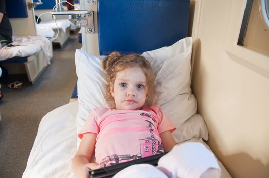 Frightened three year old girl lying on the bed with a tablet in the hands of reserved seats in the train