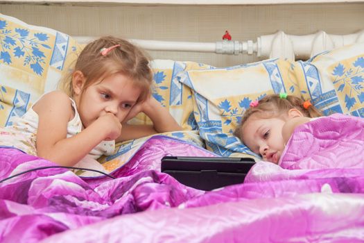 Two girls of three and five years, lie in bed and watch cartoons in the Tablet PC