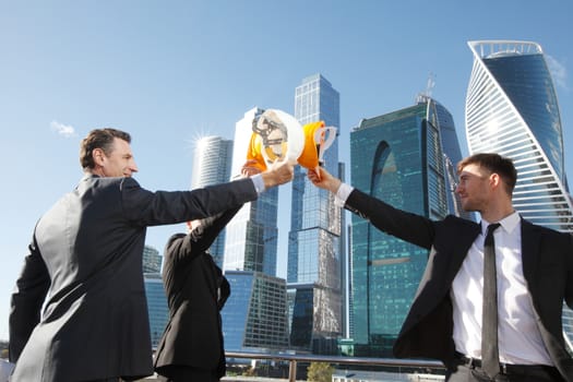 Group of business people clinking their helmets as a success on skyscrapers background, construction finished concept