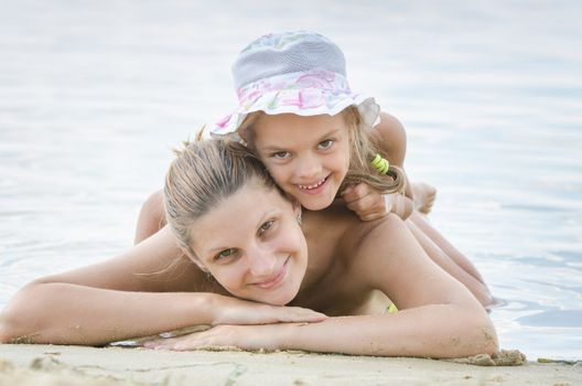 Six year old girl and a twenty-five woman lying on her stomach on the sand by the river