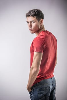 Serious looking young man wearing red t-shirt, turning around to look at camera on light background