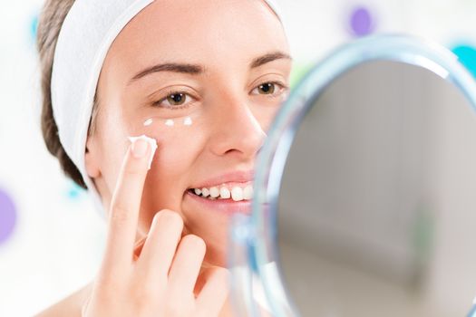 Young cute woman preparing to start her day. She is applying moisturizer cream on face. Looking at mirror and smiling. Close-up.