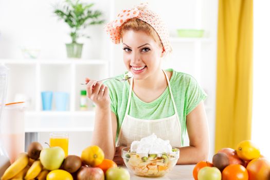 Beautiful young woman eat fruit salad with whipped cream in the kitchen. Looking at camera.