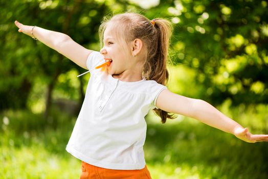 Cute Little Girl having fun in The Park and holding in mouth orange Lollipop.