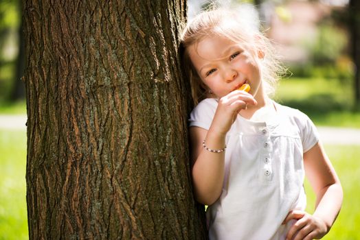 Cute Little Girl Standing in the Park leaning on a tree and holding in hand orange Lollipop.